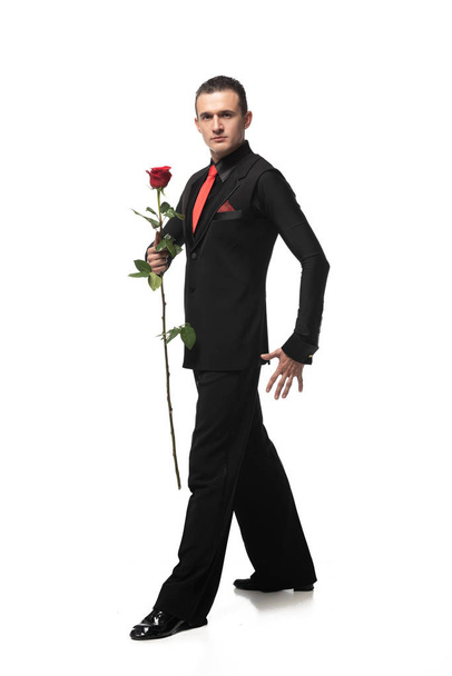 elegante tango danser in zwart pak met rode roos terwijl het kijken naar camera op witte achtergrond - Foto, afbeelding