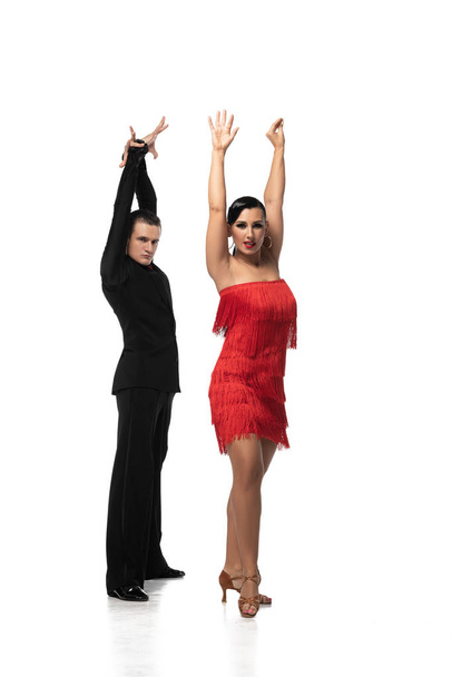 εκφραστικό ζευγάρι χορευτών που κοιτάζουν την κάμερα ενώ εκτελούν ταγκό με υψωμένα χέρια σε λευκό φόντο - Φωτογραφία, εικόνα