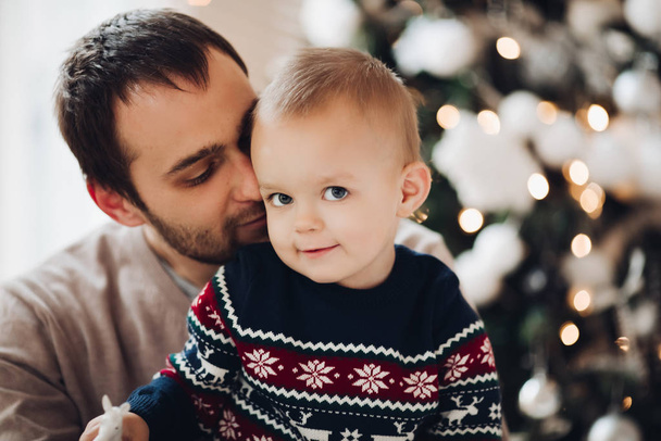 Vater küsst seinen liebenswerten Sohn gegen unkonzentrierten Weihnachtsbaum. - Foto, Bild