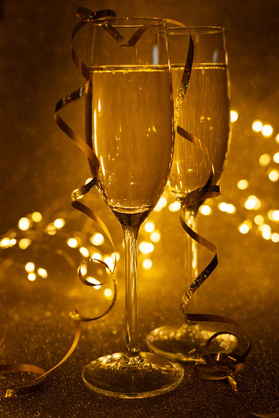 シャンパンの2つのガラスのフロントビューは、焦点を絞ったライトで金色の輝く背景にストリーマーを飾りました。クリスマスと新年の休暇の概念。スペースのコピー. - 写真・画像