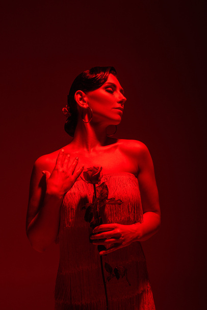 elegantní tanečnice tanga se zavřenýma očima držící červenou růži na tmavém pozadí s červeným osvětlením - Fotografie, Obrázek