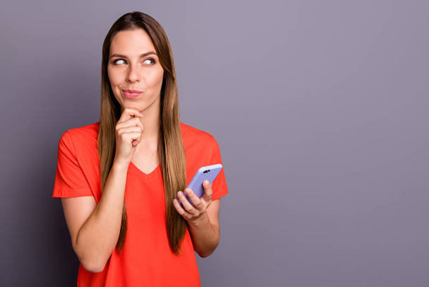 Porträt eines positiven listigen Mädchens, das süchtig nach sozialen Medien ist, schreibt SMS auf ihr Handy und denkt, Pläne berühren Kinn-Look Kopierraum tragen rotes T-Shirt isoliert über grauem Hintergrund - Foto, Bild
