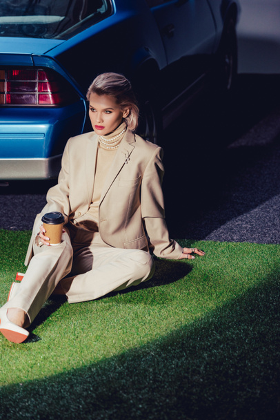attractive and stylish woman in suit sitting near retro car and holding paper cup  - Foto, Imagem