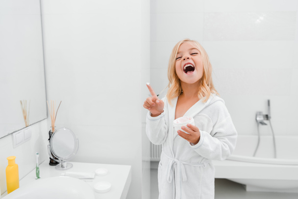 szczęśliwy dziecko z kremem kosmetycznym na palec śmiejąc się w łazience - Zdjęcie, obraz