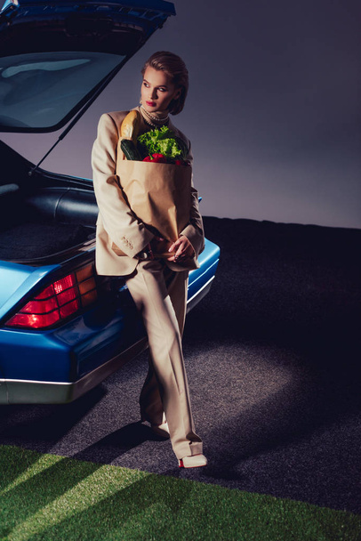 attractive and stylish woman in suit standing near retro car and holding paper bag with food  - Фото, зображення