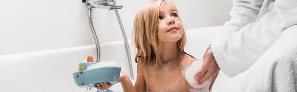 Panoramaaufnahme eines süßen Kindes, das Gummispielzeug in der Hand hält und Mutter im Badezimmer ansieht  - Foto, Bild