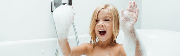 Panoramaaufnahme eines Kindes mit geöffnetem Mund und Badeschaum an den Händen  - Foto, Bild