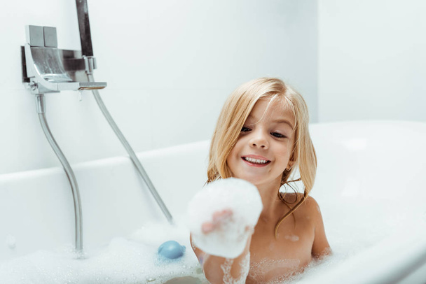 szczęśliwy nagi dzieciak patrząc na pianę kąpielową na rękę podczas kąpieli  - Zdjęcie, obraz