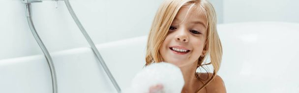 panoramic shot of happy naked kid looking at bath foam on hand while taking bath  - Фото, изображение