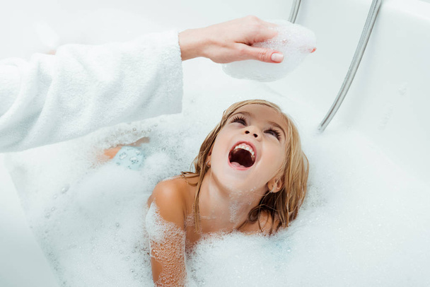 happy kid looking at hand of mother in bath foam in bathroom  - Foto, afbeelding