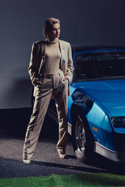 attractive and stylish woman in suit with hands in pockets standing near retro car  - Foto, Imagen