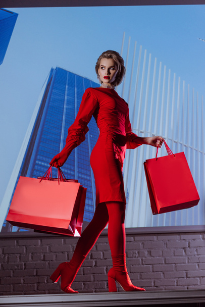 Blick auf eine attraktive und stilvolle Frau im roten Kleid, die Einkaufstüten vor dem Hintergrund der Stadt hält  - Foto, Bild
