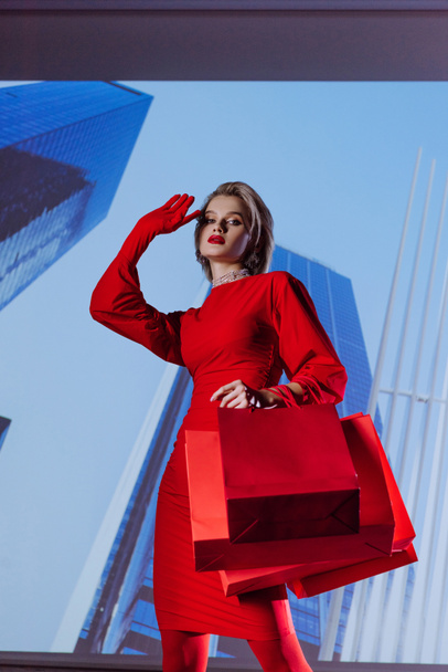low angle view of attractive and stylish woman in red dress holding shopping bags on city background  - Valokuva, kuva