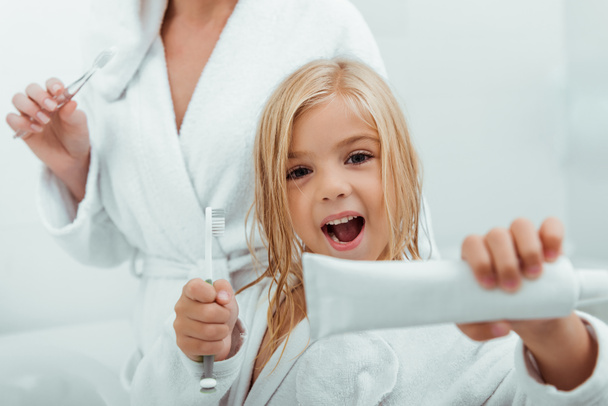 enfoque selectivo de niño alegre sosteniendo pasta de dientes y cepillo de dientes cerca de la madre
  - Foto, Imagen