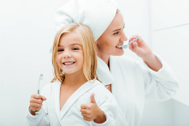 cute child holding toothbrush and showing thumb up near mother  - Foto, Imagem