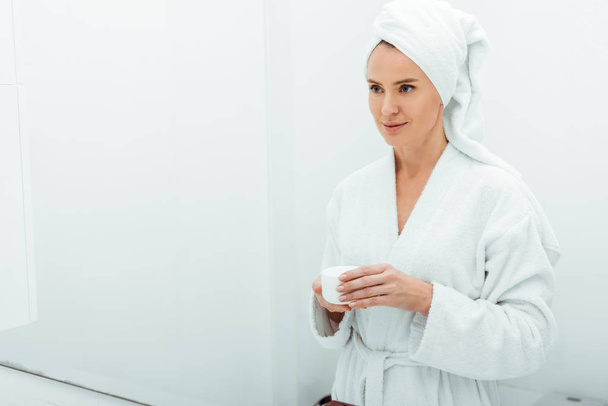 bella donna in accappatoio e asciugamano contenitore con crema viso in bagno
  - Foto, immagini