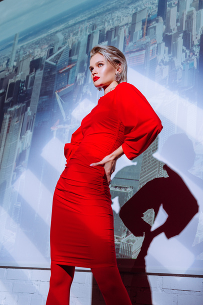 low angle view of attractive and stylish woman with hand on hip in red dress on city background  - Foto, Imagem