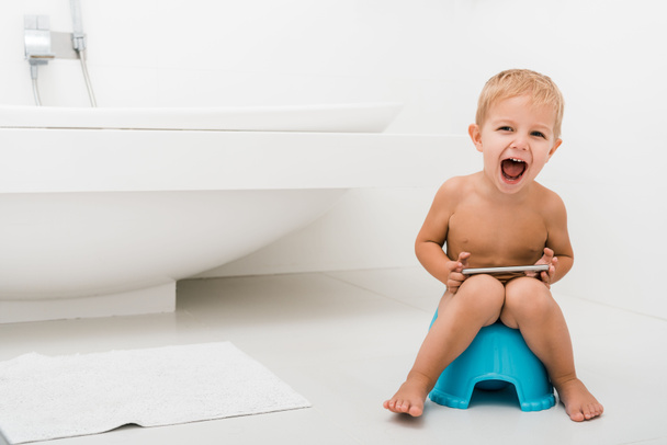 aufgeregter Kleinkind Junge mit geöffnetem Mund sitzt auf blauem Töpfchen und hält Smartphone in der Nähe der Badewanne  - Foto, Bild