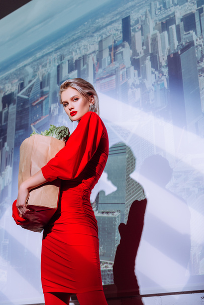 Blick auf eine attraktive und stilvolle Frau in rotem Kleid, die eine Papiertüte mit Lebensmitteln vor dem Hintergrund der Stadt hält  - Foto, Bild