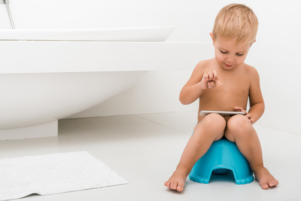 cute toddler boy sitting on blue potty and using smartphone near bathtub  - Foto, Imagen