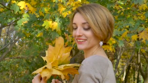 Emozionale attraente giovane donna con capelli corti biondi e vestito da biege che tiene un mazzo di foglie autunnali sopra la testa e posa in un bellissimo parco all'aperto
 - Filmati, video