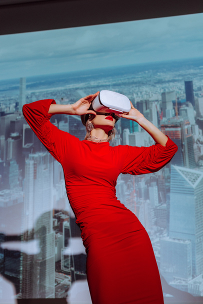 άποψη χαμηλή γωνία της κομψής γυναίκας σε κόκκινο φόρεμα και εικονικής πραγματικότητας ακουστικά στο φόντο της πόλης  - Φωτογραφία, εικόνα