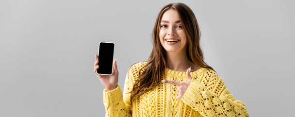 πανοραμική λήψη χαρούμενης νεαρής γυναίκας που δείχνει με το δάχτυλο το smartphone με μαύρη οθόνη απομονωμένη σε γκρι - Φωτογραφία, εικόνα