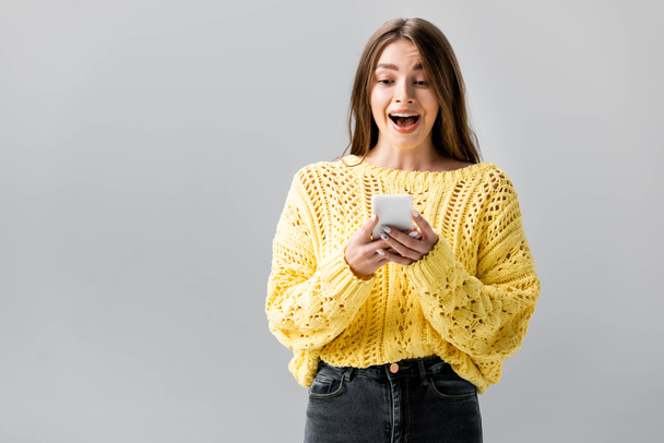 surprised girl in yellow sweater looking at smartphone isolated on grey - Fotoğraf, Görsel