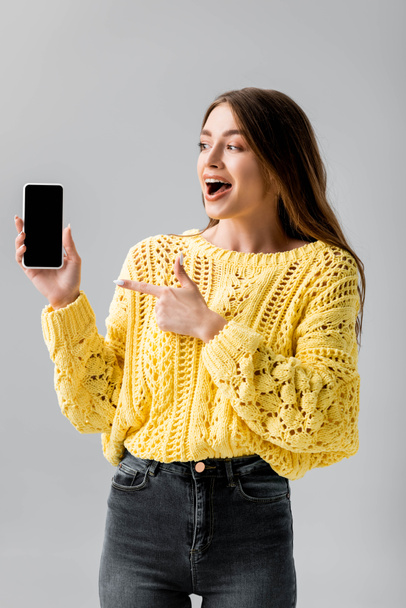 cheerful young woman pointing with finger at smartphone with blank screen isolated on grey - Photo, Image
