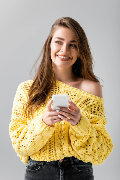 smiling girl in yellow sweater looking away while using smartphone isolated on grey - Foto, imagen