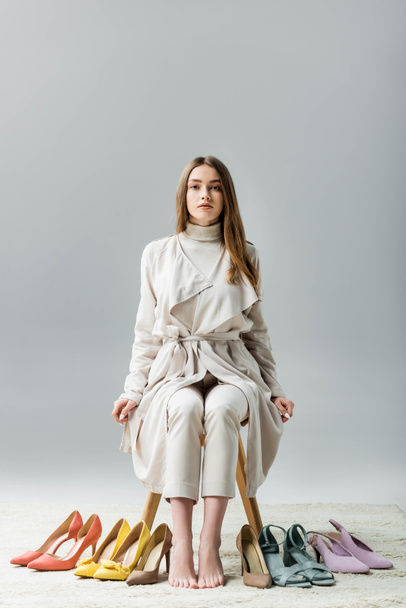 stylish, confident girl looking at camera while sitting on chair near collection of shoes on grey background - Фото, изображение