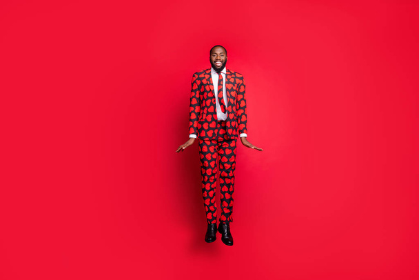 Full size photo of crazy dark skin hipster guy jumping high demonstrating cool modern trendy look wear stylish hearts pattern suit blazer shirt necktie tie trousers isolated red color background - 写真・画像