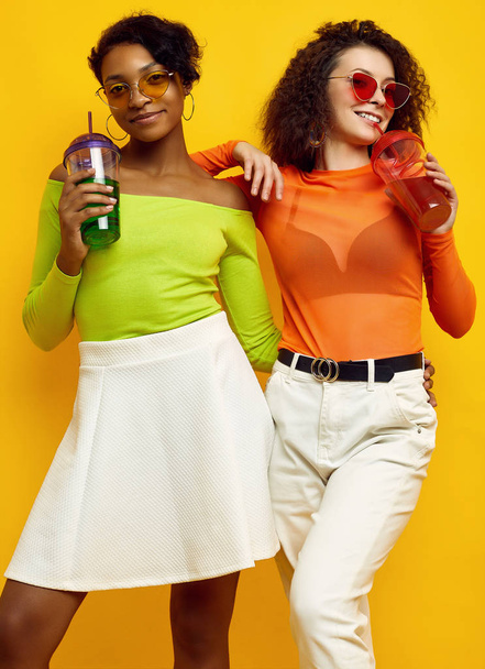Two young beautiful hipster girls in colorful summer clothes with cocktails - 写真・画像