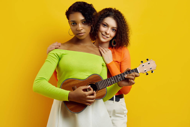 Two young beautiful hipster girls in colorful summer clothes with small guitar - Foto, Imagen