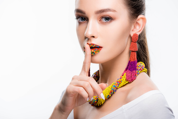 attractive girl in beaded accessories, with beads on lips, showing hush sign and looking at camera isolated on white - Photo, Image