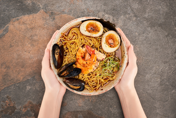 cropped view of woman holding bowl with spicy seafood ramen on stone surface - Foto, Imagem