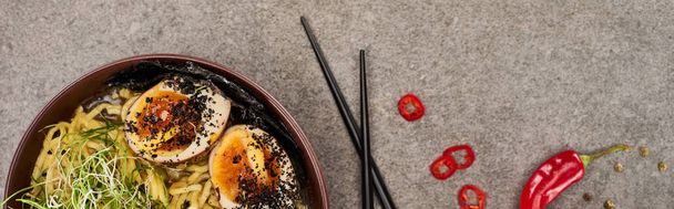 top view of spicy meat ramen near chili pepper and chopsticks on grey concrete surface, panoramic shot - Foto, Bild