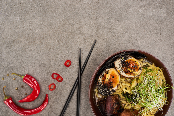 top view of spicy meat ramen near chili pepper and chopsticks on grey concrete surface - Photo, Image