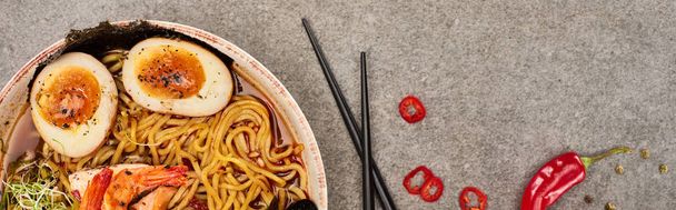 top view of spicy seafood ramen near chili pepper and chopsticks on grey concrete surface, panoramic shot - Foto, Imagem