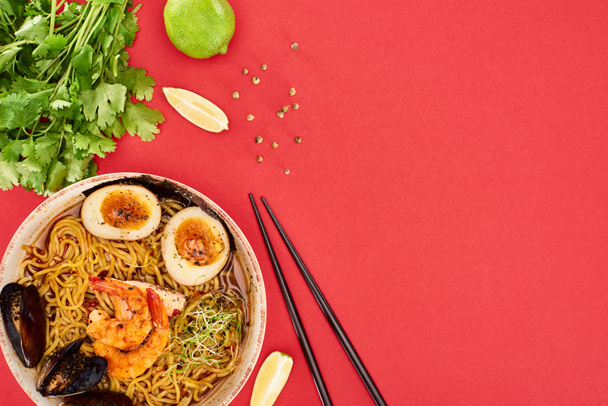 top view of spicy seafood ramen near chopsticks, parsley and lime isolated on red - Фото, изображение