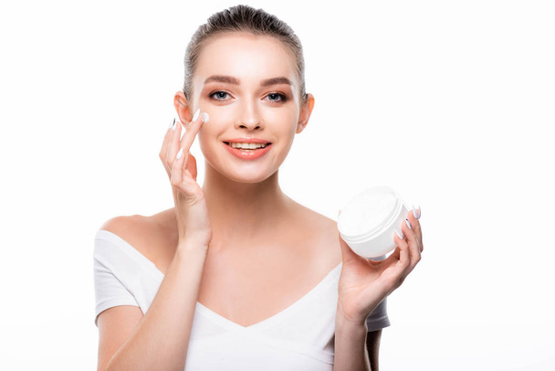 attractive, smiling woman applying face cream and looking at camera isolated on white - Фото, изображение