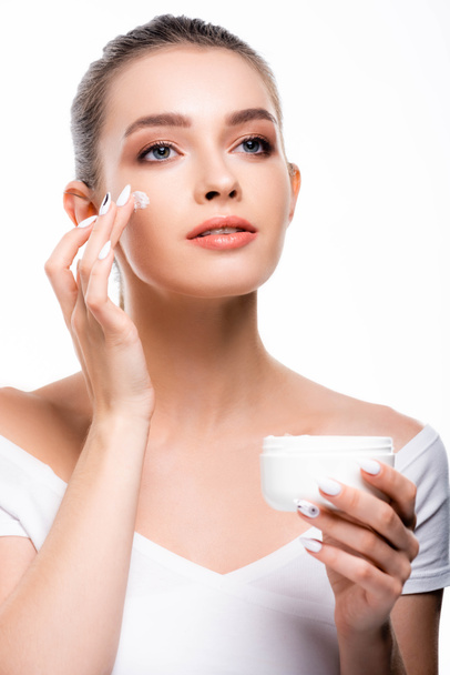 beautiful, smiling woman applying cosmetic cream on face and looking away isolated on white - Zdjęcie, obraz