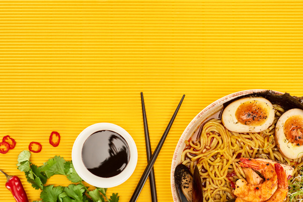 top view of seafood ramen near fresh ingredients, soy sauce and chopsticks on yellow surface - Фото, зображення