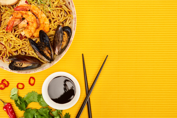 top view of seafood ramen near fresh ingredients, soy sauce and chopsticks on yellow surface - Photo, Image