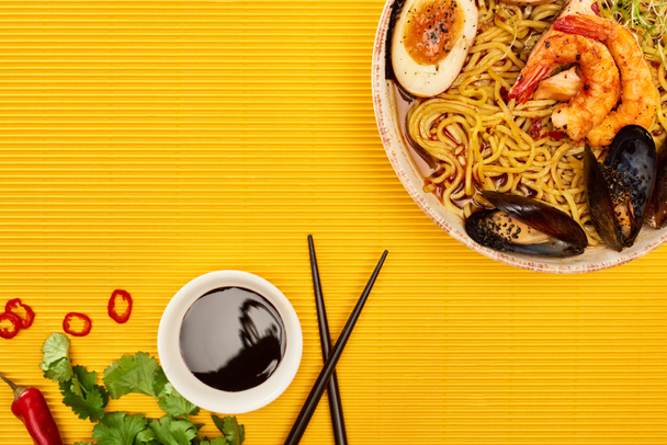 top view of seafood ramen near fresh ingredients, soy sauce and chopsticks on yellow surface - Foto, Imagen