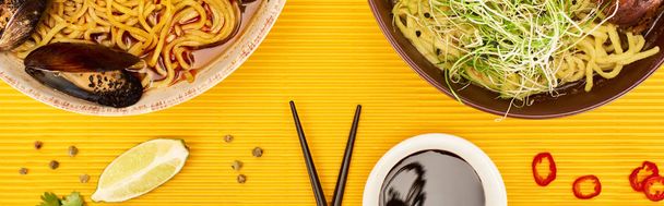 top view of seafood and meat ramen near fresh ingredients, soy sauce and chopsticks on yellow surface, panoramic shot - Foto, Bild