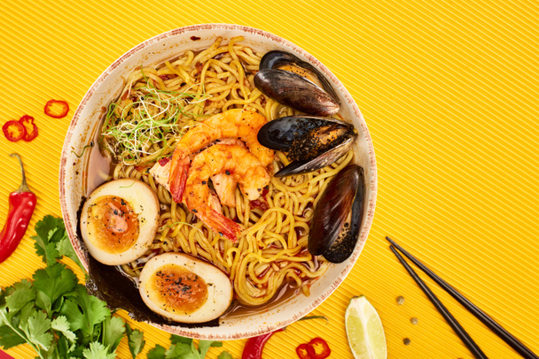 top view of seafood ramen near fresh ingredients and chopsticks on yellow surface - Valokuva, kuva