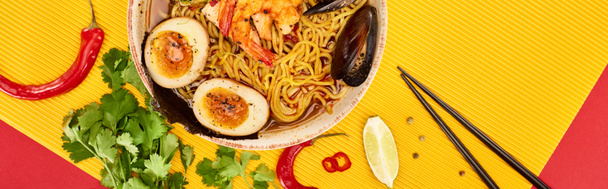top view of seafood ramen near fresh ingredients and chopsticks on yellow and red surface, panoramic shot - Foto, Bild