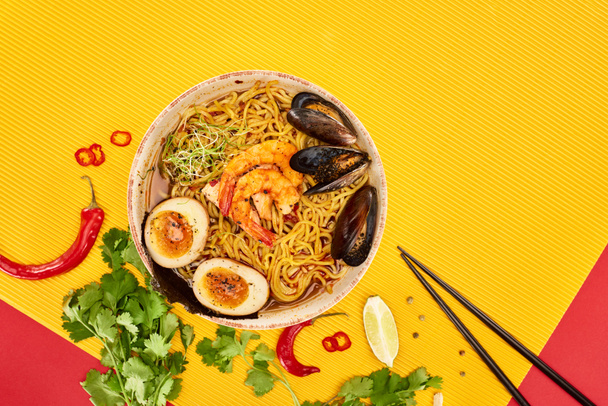 top view of seafood ramen near fresh ingredients and chopsticks on yellow and red surface - Photo, Image