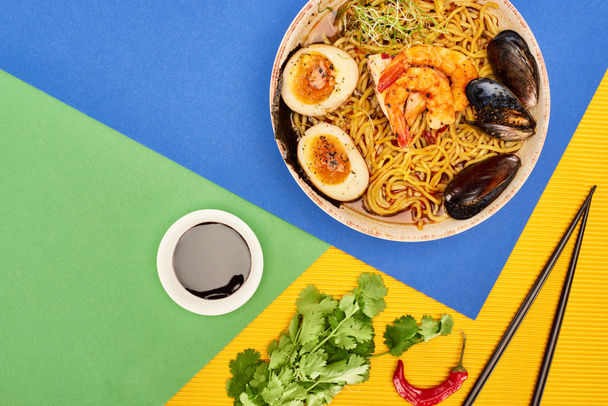 top view πικάντικα θαλασσινά ράμεν κοντά σε φρέσκα υλικά, σάλτσα τσίλι και chopsticks στην πολύχρωμη επιφάνεια - Φωτογραφία, εικόνα
